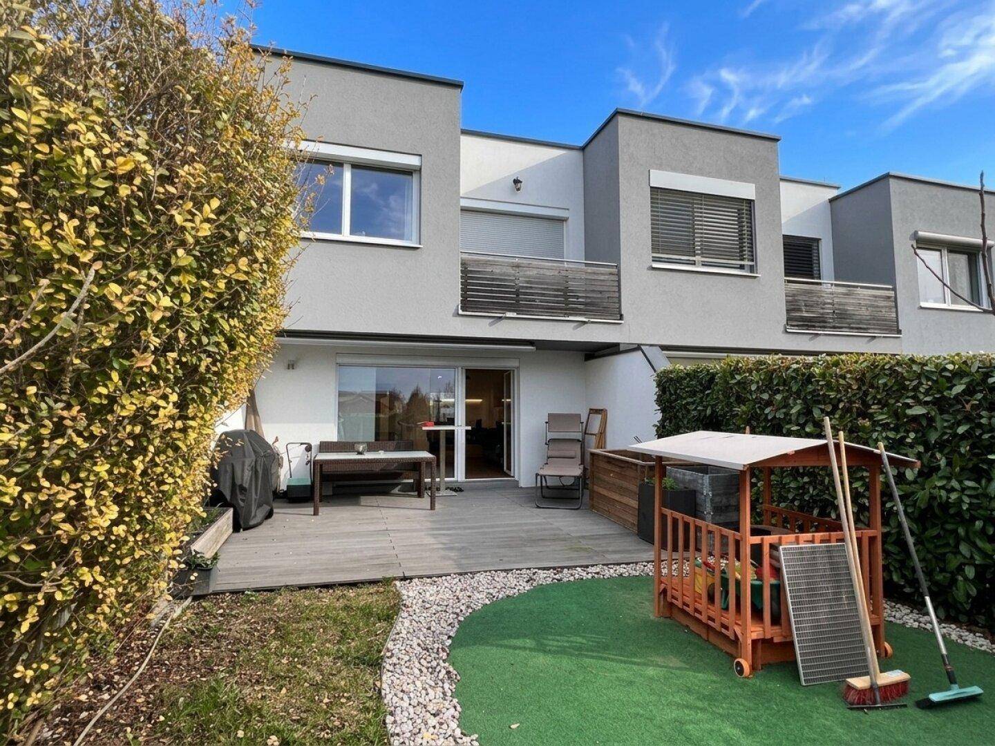 Moderne Reihenhausfassade mit Holzterrasse, Gartenmöbeln und einem kleinen Spielbereich im Grünen.