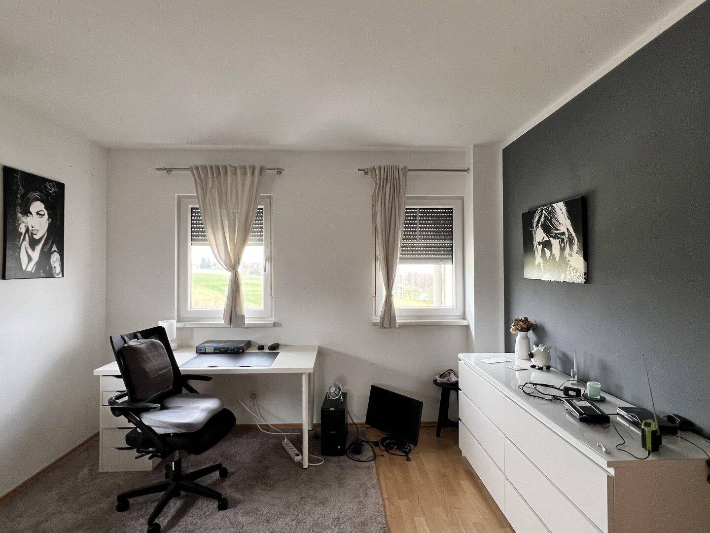 Arbeitszimmer mit Schreibtisch, grauem Teppich und Blick ins Grüne durch die Fenster.
