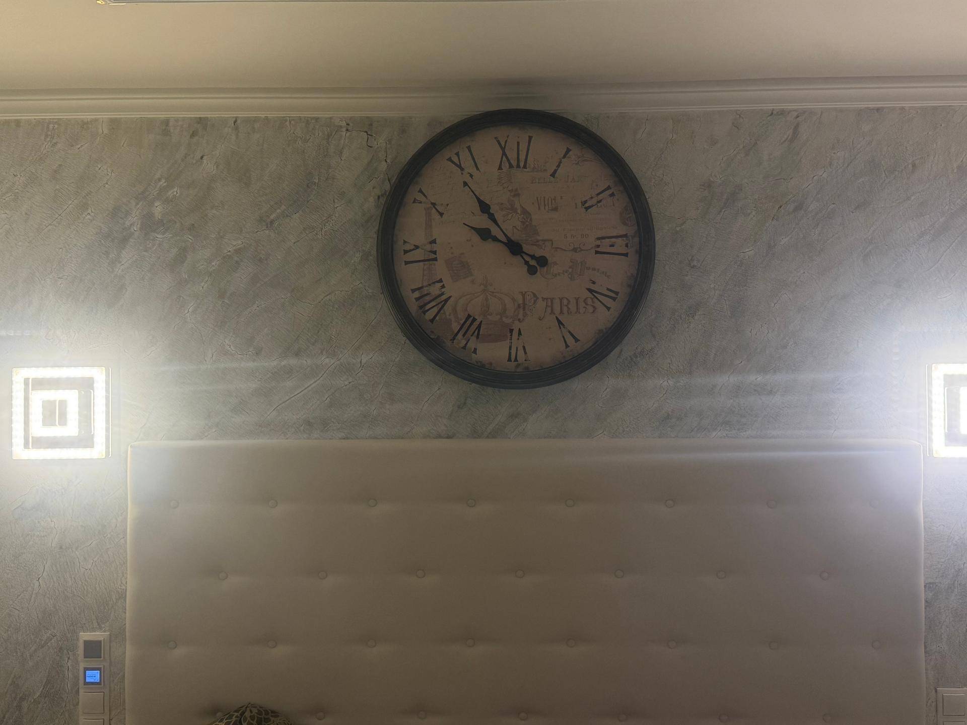 Schlafzimmerwand mit großem Zifferblatt und gepolstertem Kopfteil, akzentuiert durch moderne Wandleuchten.