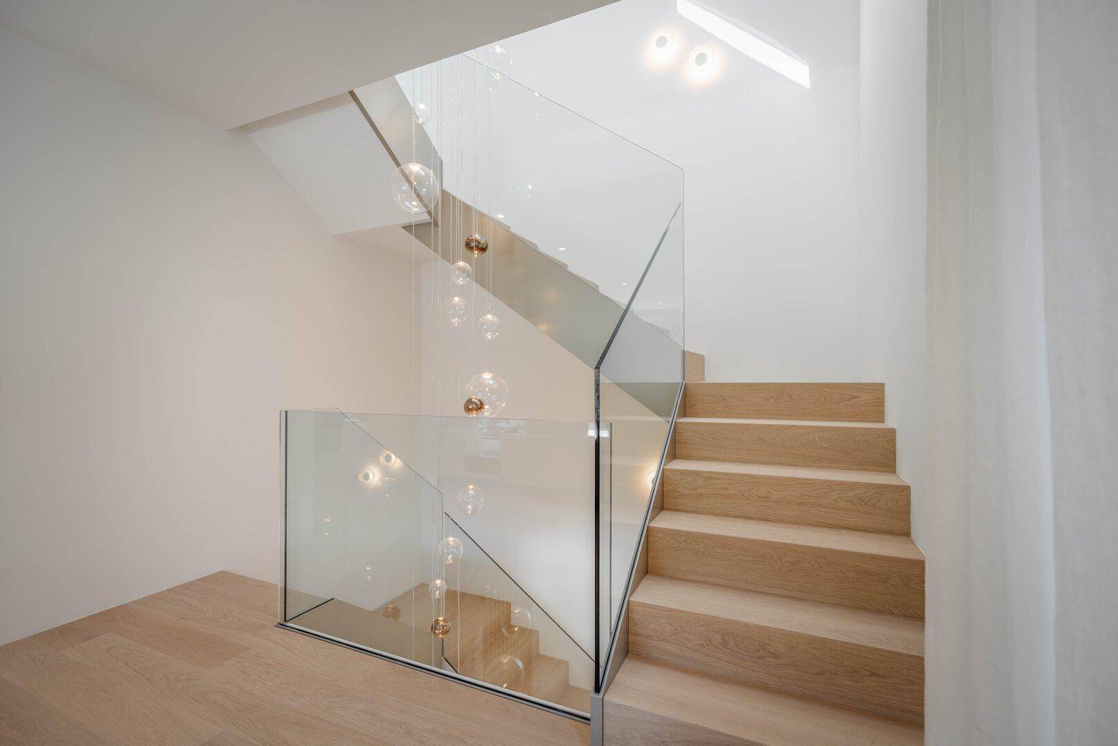 Moderne Treppe mit Glasgeländer und schwebenden Leuchten, die den Raum erhellen.