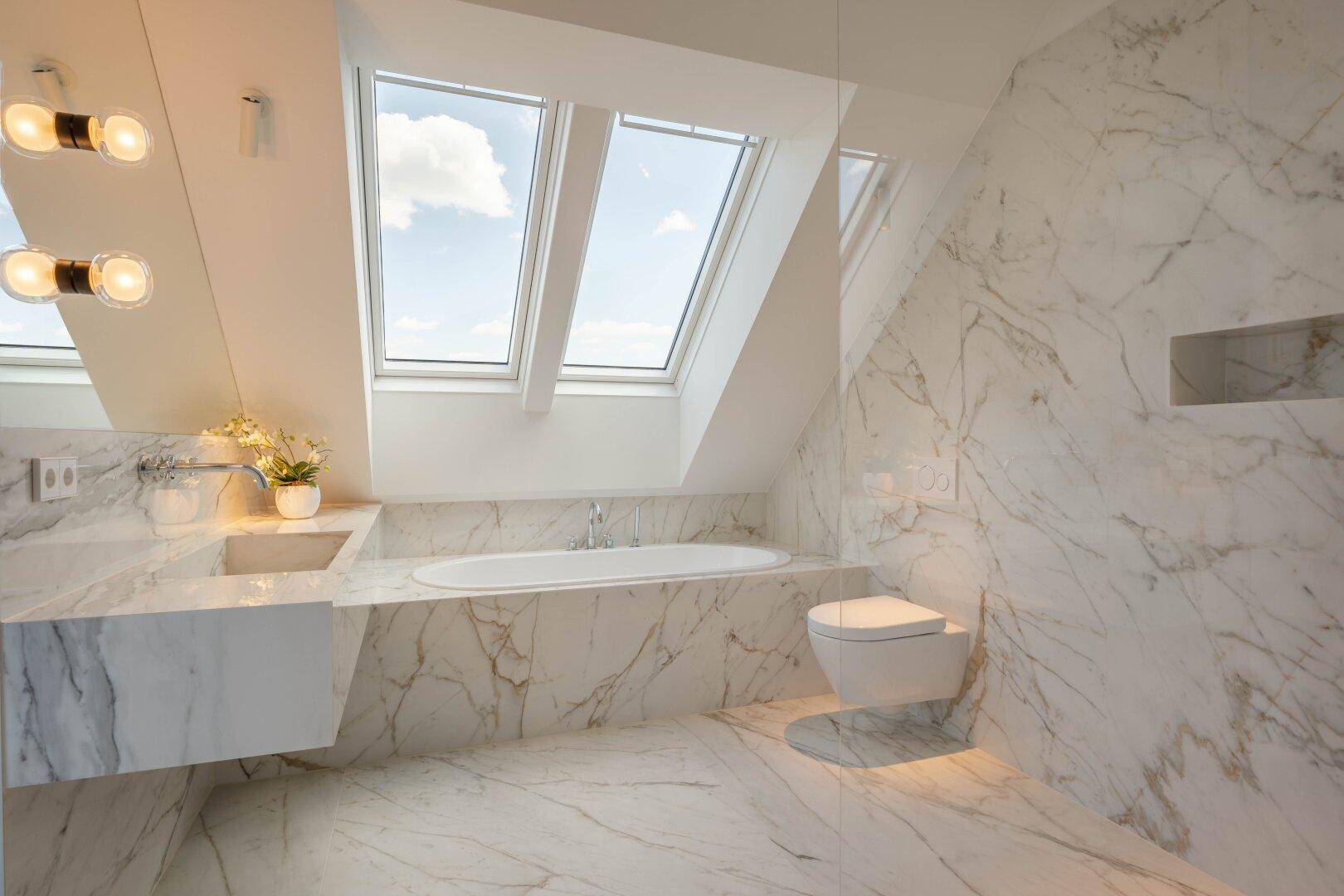 Elegantes Badezimmer mit Marmoroberflächen, Badewanne, wandmontierter Toilette und Dachfenstern.