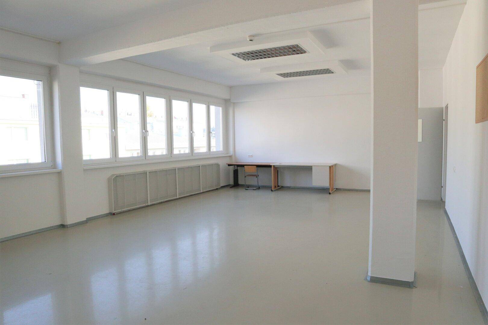 Heller, leerer Raum mit großen Fenstern, Heizkörpern und funktionaler Deckenbeleuchtung, bereit zur Einrichtung.