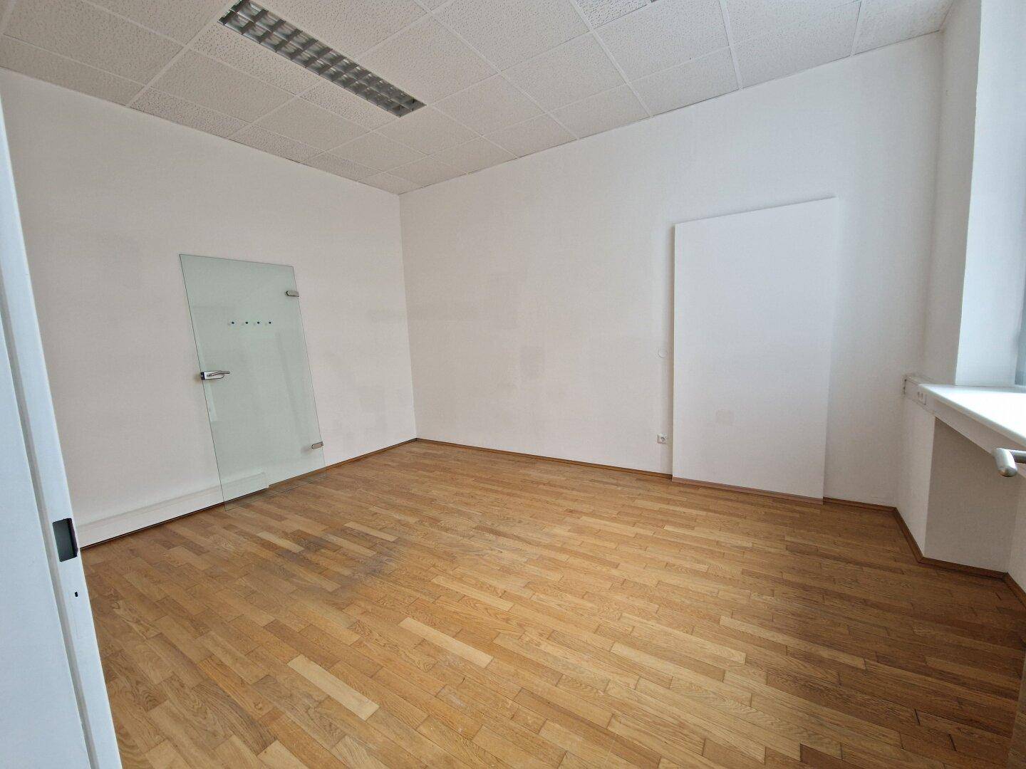 Leerer Raum mit Holzparkettboden und einer modernen Glastür, geeignet für verschiedene Nutzungen.