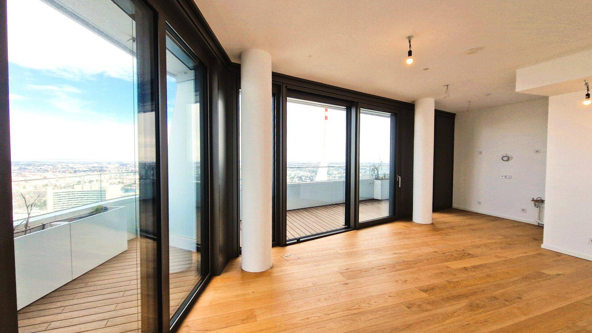 Heller Wohnraum mit großen Fensterfronten, Holzboden und Zugang zu einem Balkon.