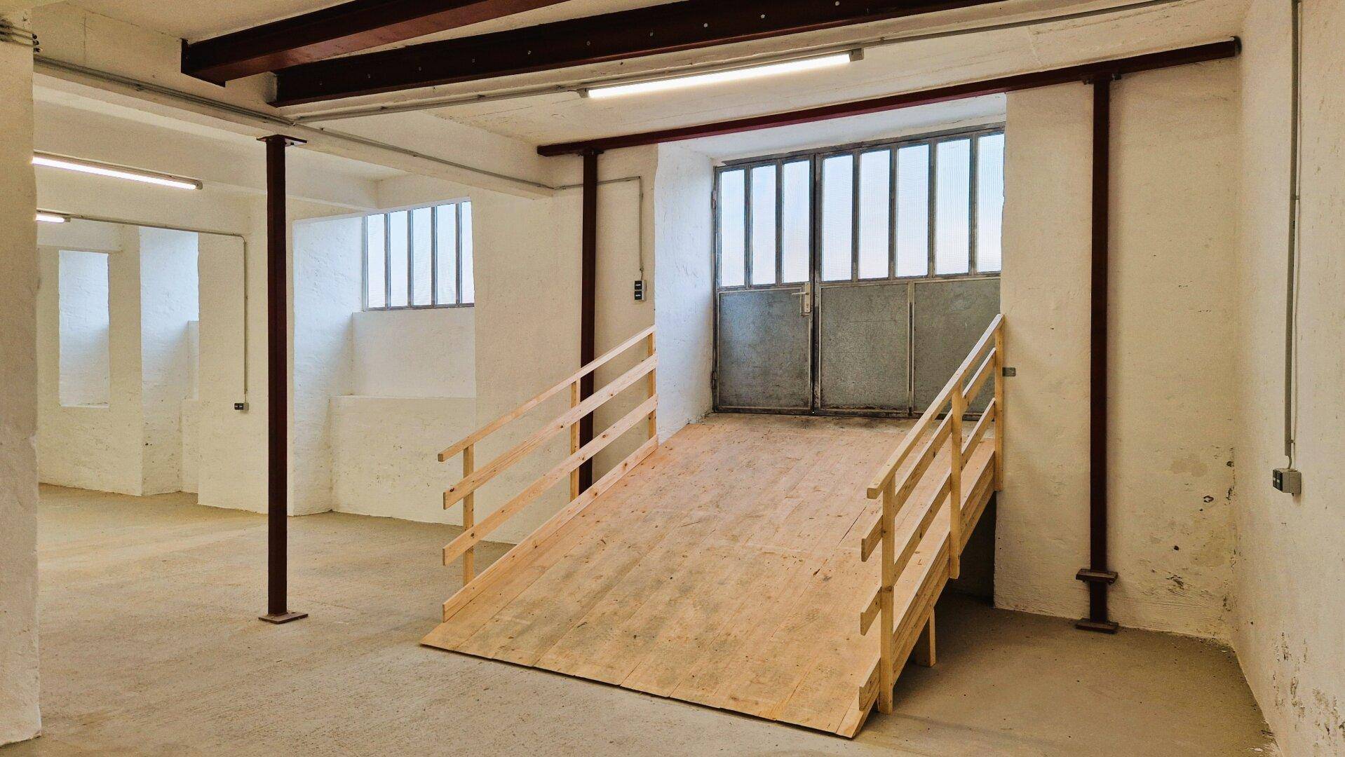 Großer Lagerraum mit einer Holzrampe, die zu breiten Doppeltüren führt, Betonboden und sichtbaren Stahlträgern.