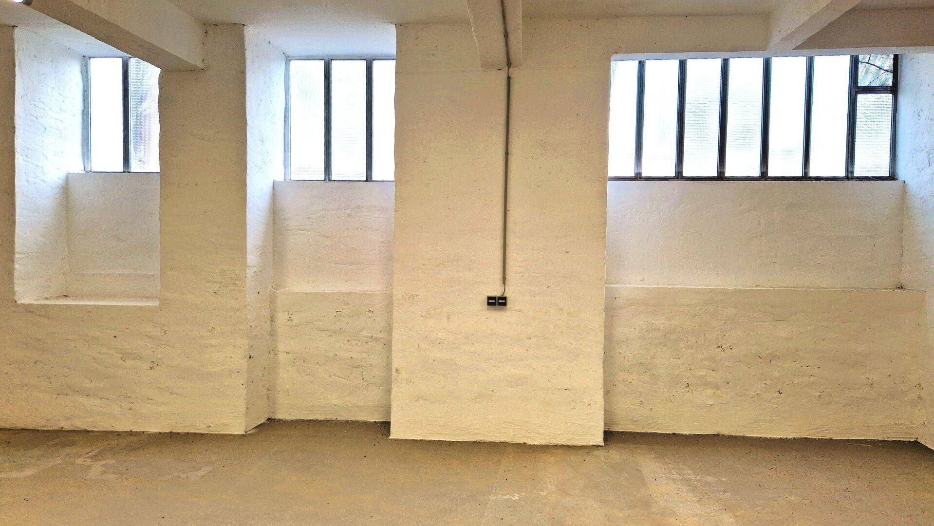 Detailansicht einer weißen Wand in einem Lagerraum mit Betonboden und mehreren kleinen, vergitterten Fenstern.