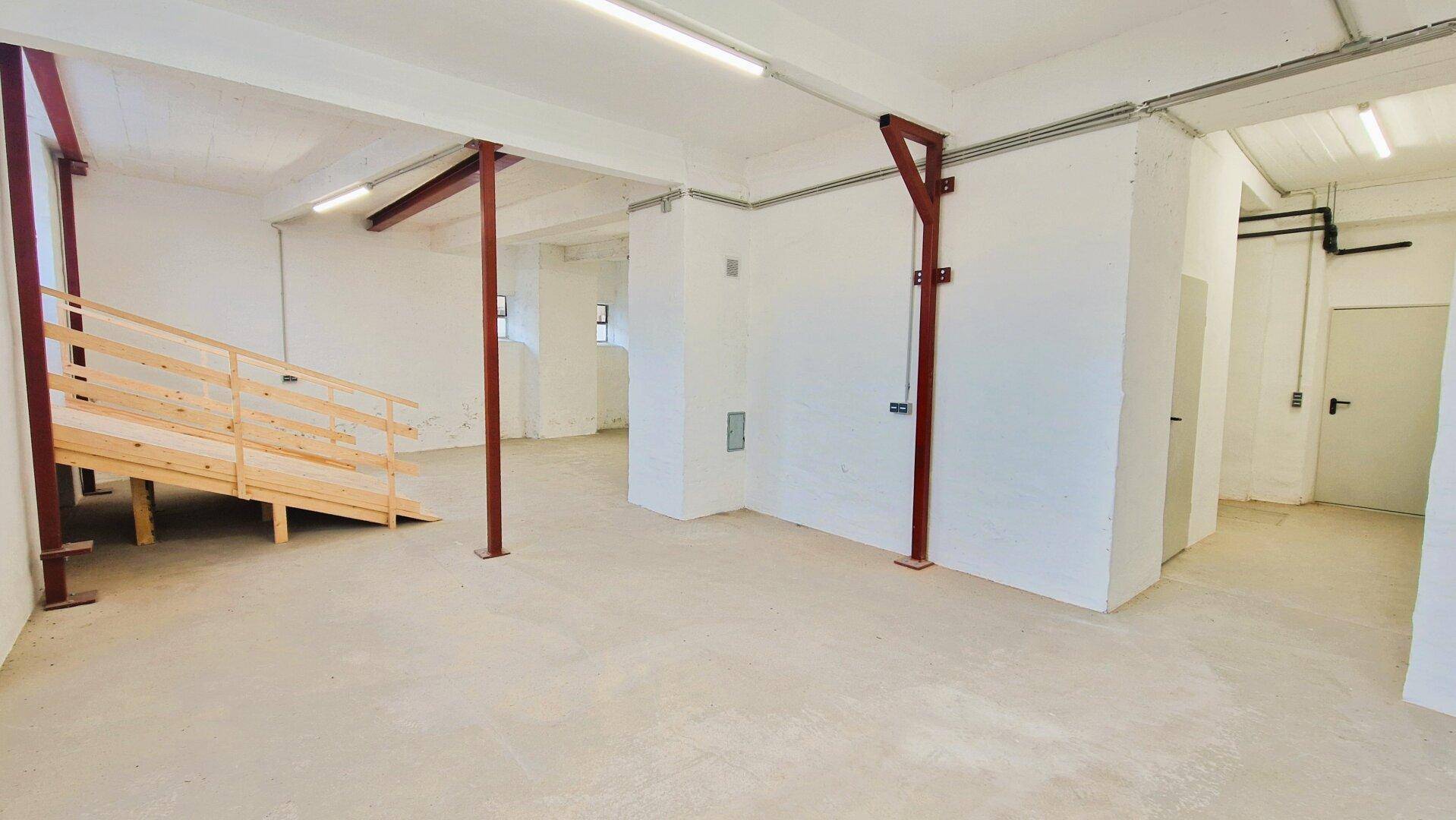 Heller Lagerraum mit einer Holzrampe, Betonboden, hohen Decken und sichtbaren roten Stahlträgern.