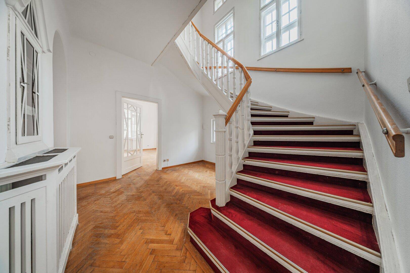 Elegantes Treppenhaus mit rotem Teppich, Holzgeländer und natürlichem Lichteinfall.