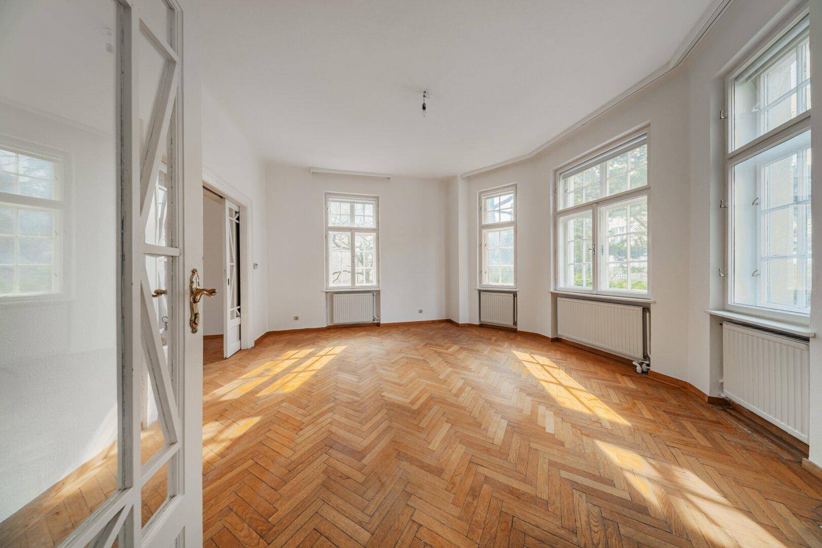 Heller Wohnraum mit Fischgrätparkett, großen Fenstern und viel Sonnenlicht.