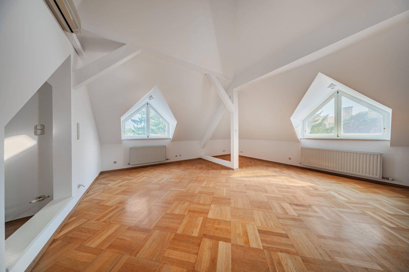 Ausgebauter Dachbodenraum mit schrägen Decken, Parkettboden und dreieckigen Fenstern.