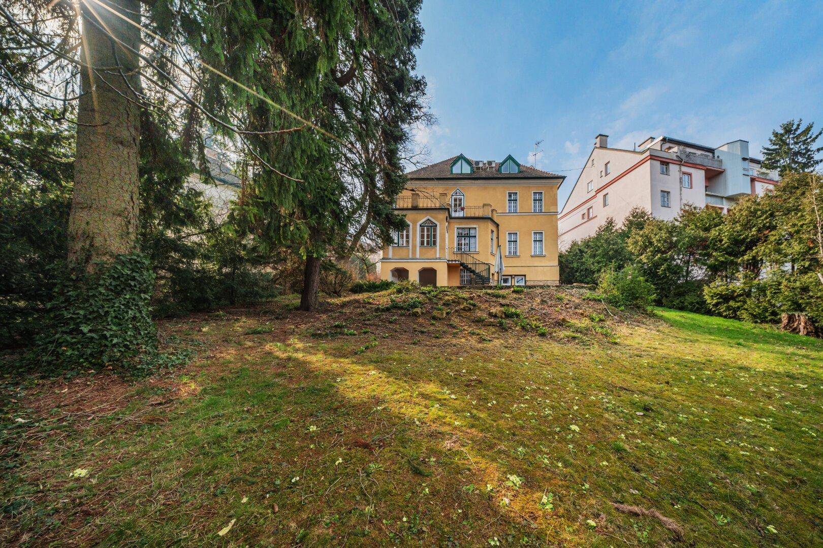 Historisches Gebäude eingebettet in einen weitläufigen Garten mit altem Baumbestand und Rasenflächen.