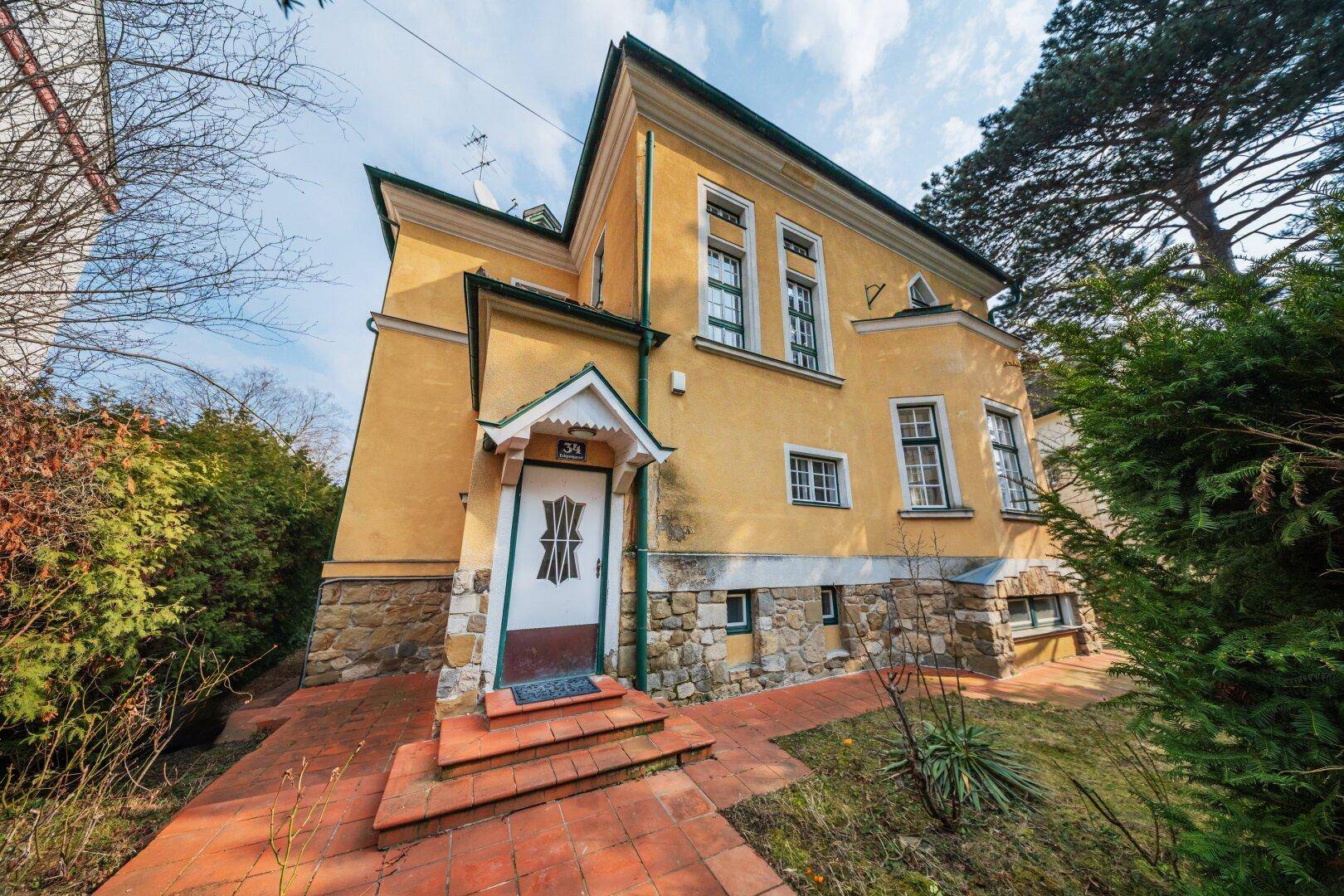 Seitenansicht des historischen Gebäudes mit Eingangsbereich und Steinsockel in grüner Umgebung.