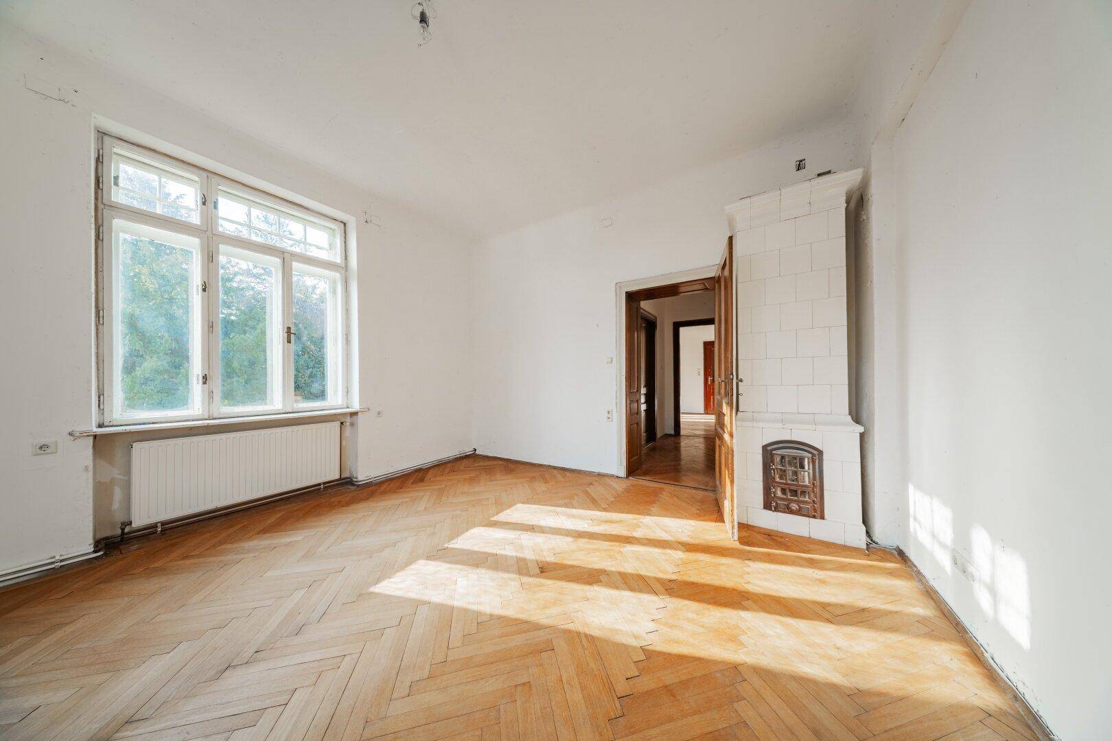 Ein helles Schlafzimmer mit Fischgrätparkett, einem großen Fenster und einem traditionellen Kachelofen.