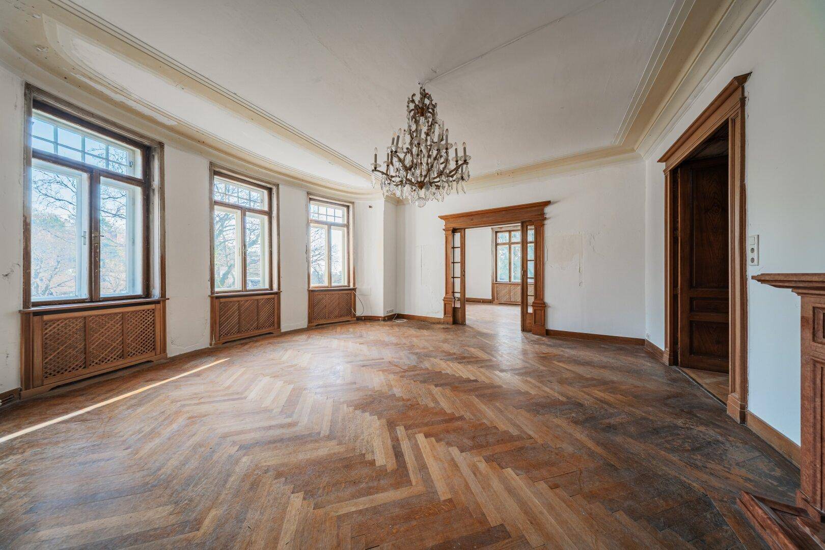 Ein geräumiges Wohnzimmer mit Fischgrätparkett, hohen Decken und einem prächtigen Kronleuchter.
