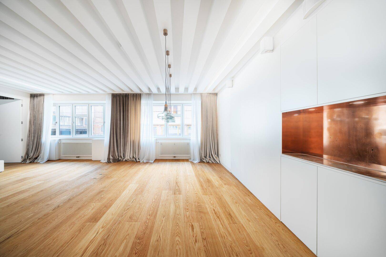 Geräumiges Wohnzimmer mit Holzbalkendecke, Parkettboden und zwei großen Fenstern.