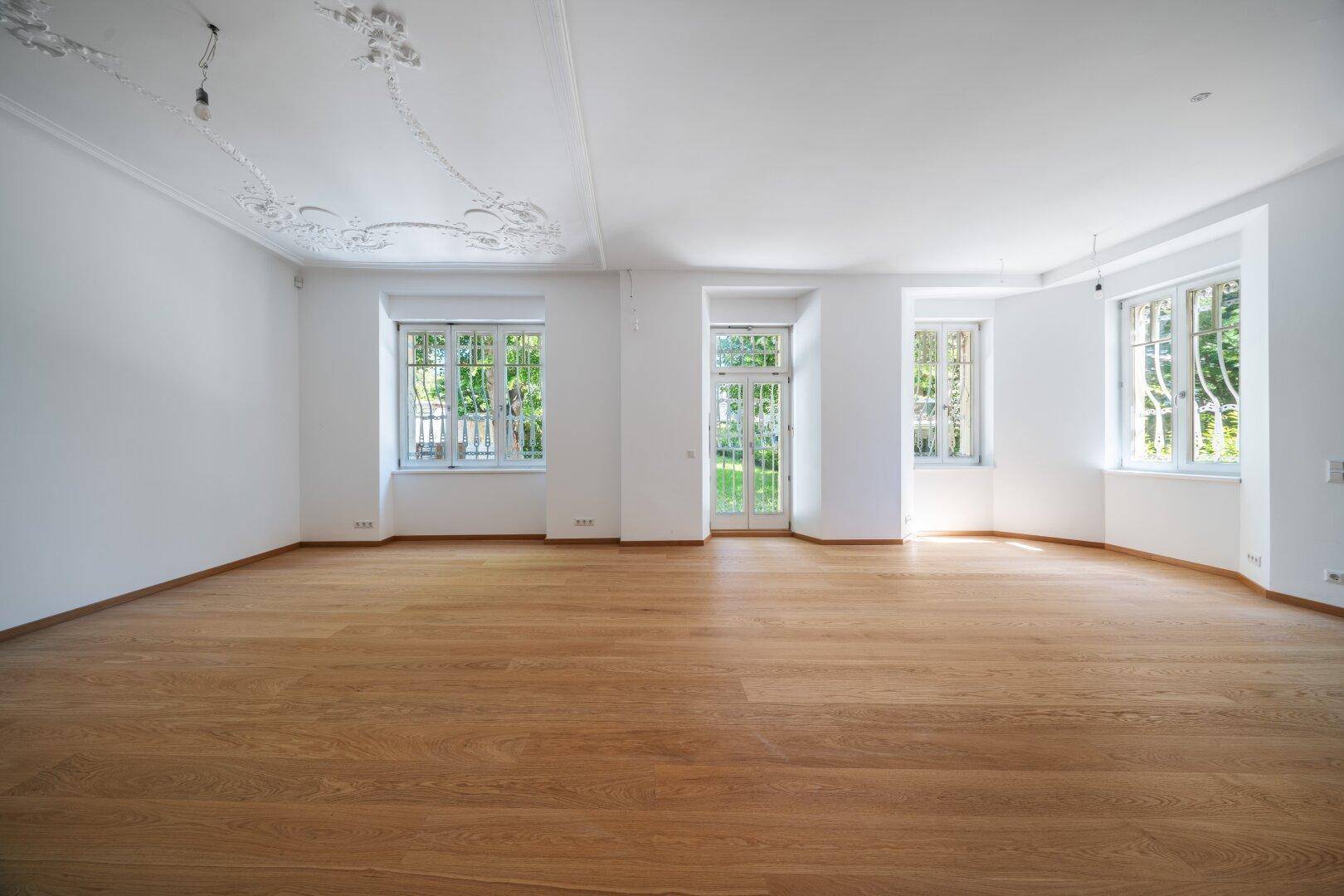 Helles, leeres Zimmer mit Eichenparkett, Stuckdecke und Blick in den Garten.