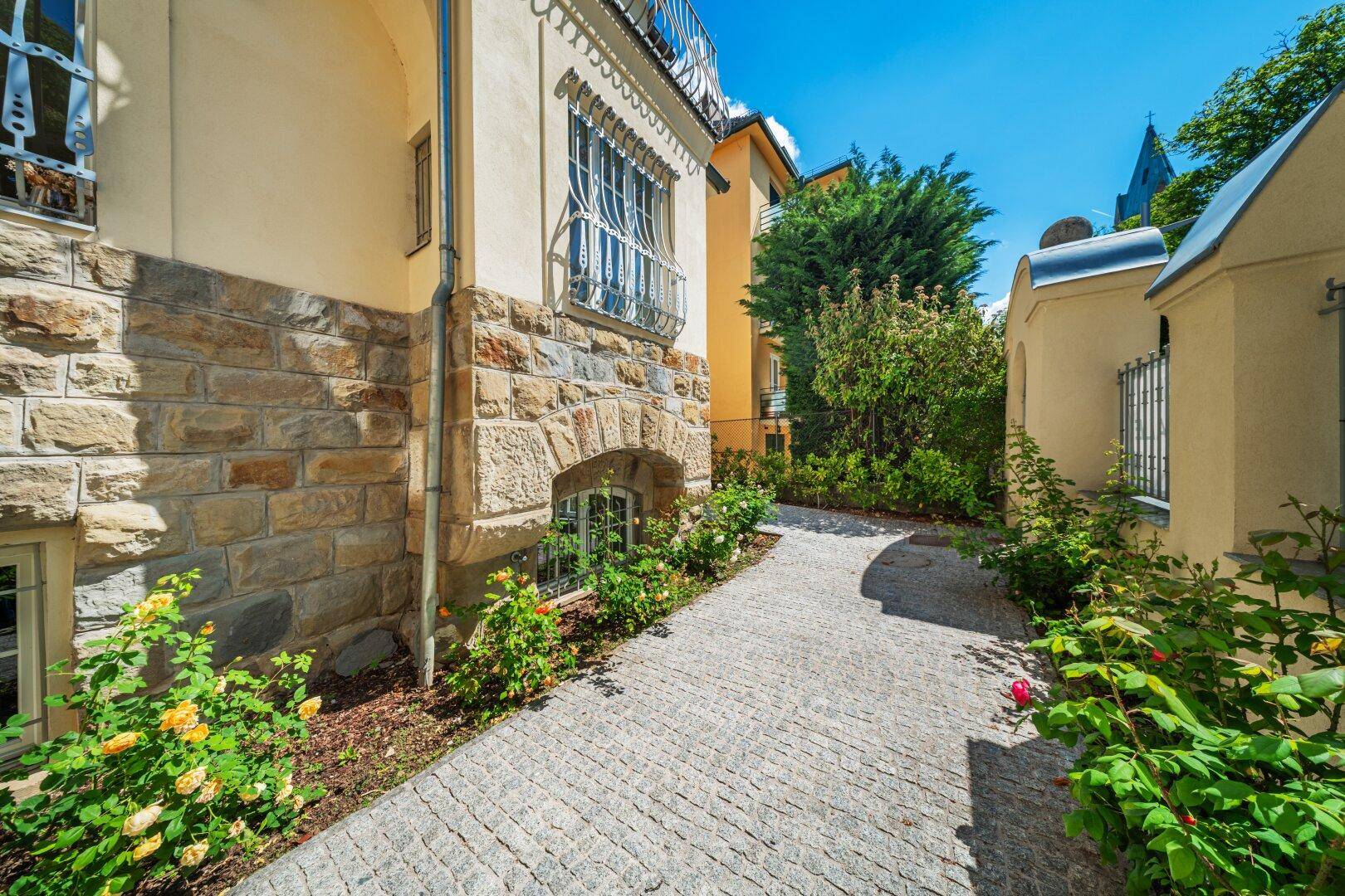 Seitenansicht der Stadtvilla mit Natursteinsockel, gepflastertem Weg und üppiger Bepflanzung.