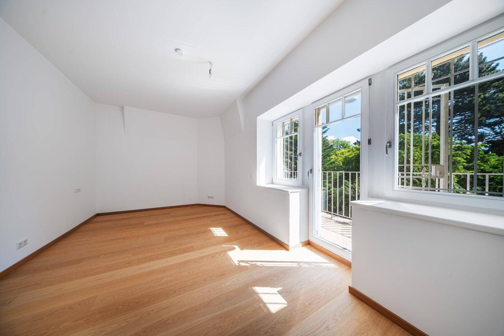 Helles, leeres Zimmer mit Holzboden, großen Fenstern und Zugang zu einem französischen Balkon.