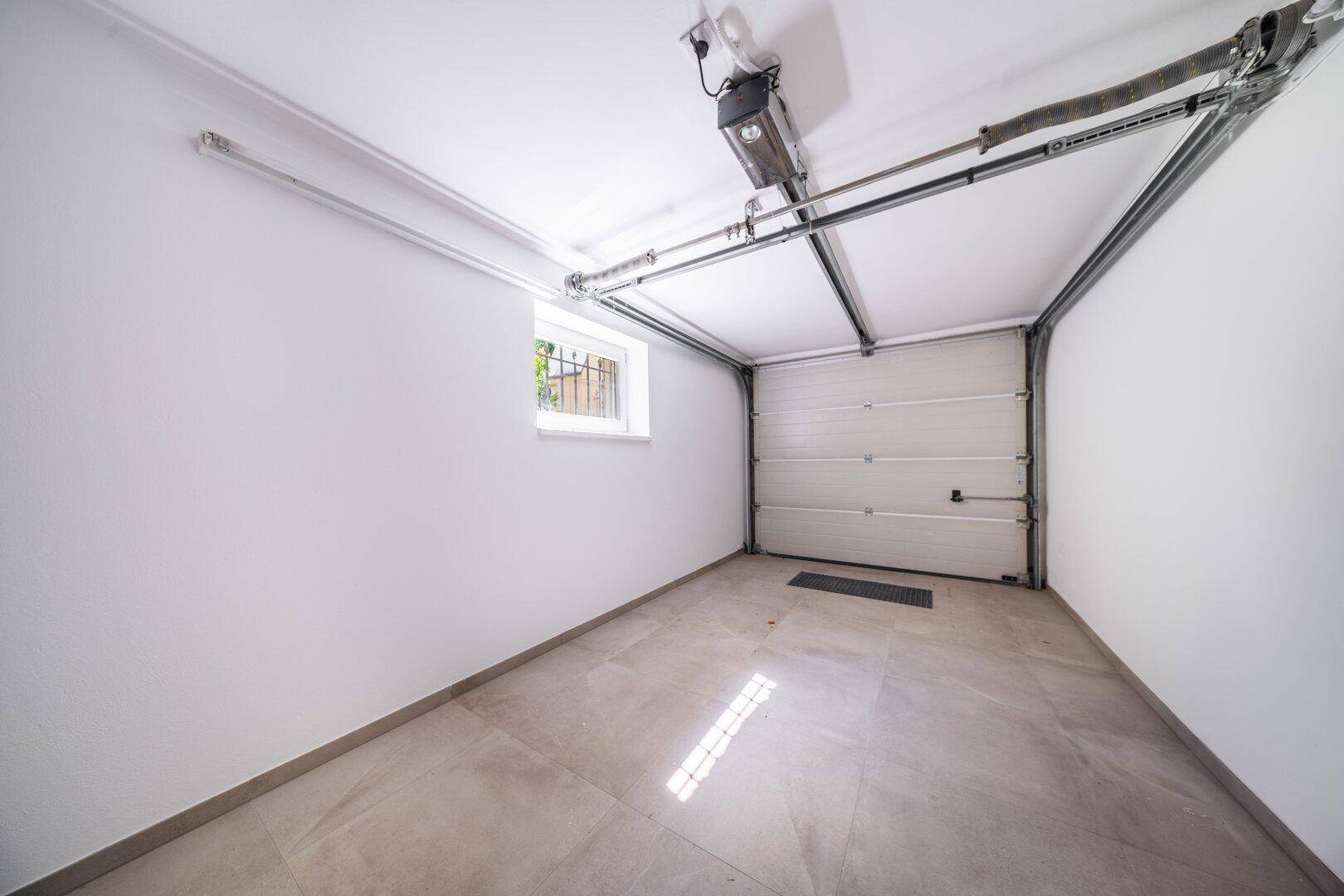 Saubere, leere Garage mit Fliesenboden, Fenster und automatischem Garagentor.