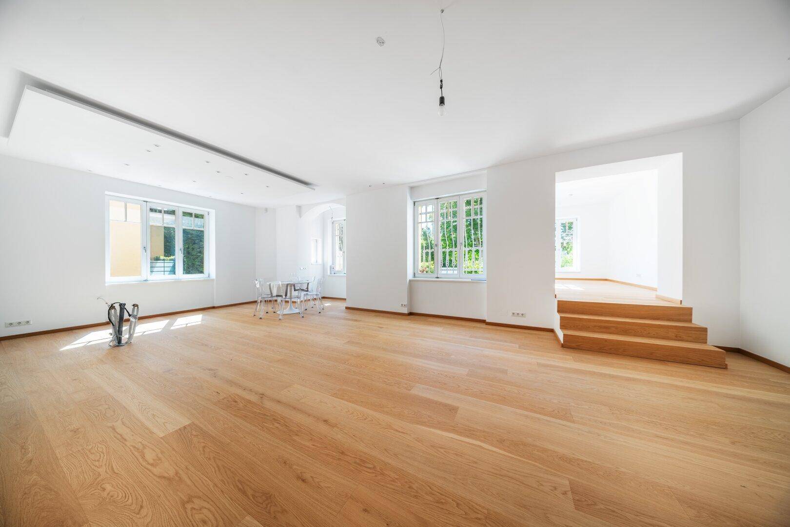 Weitläufiger Wohnbereich mit Holzboden, Stufen zu einem weiteren Bereich und großen Fenstern.