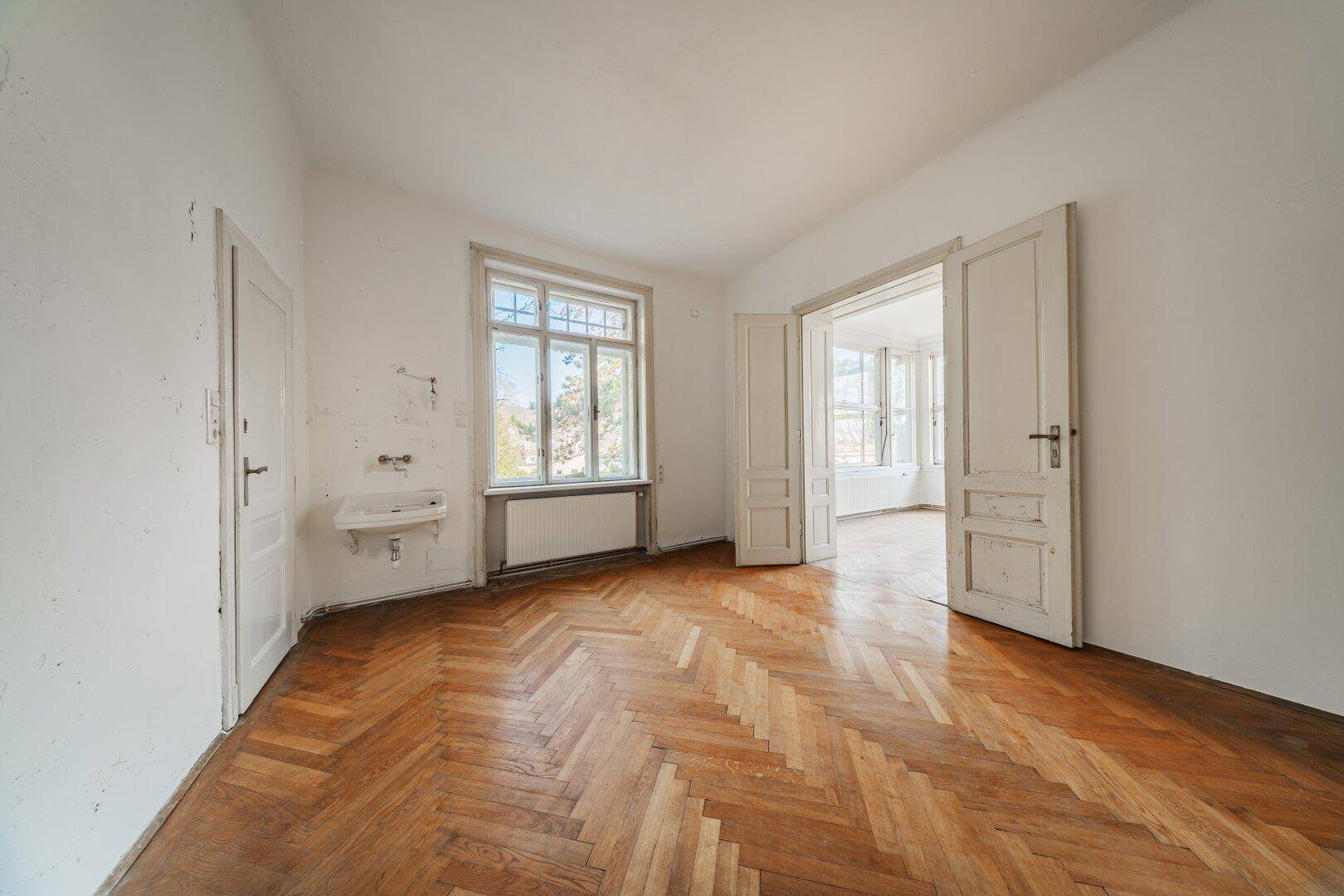 Ein leeres Zimmer mit Fischgrätparkett, einem Fenster und einem kleinen Waschbecken.