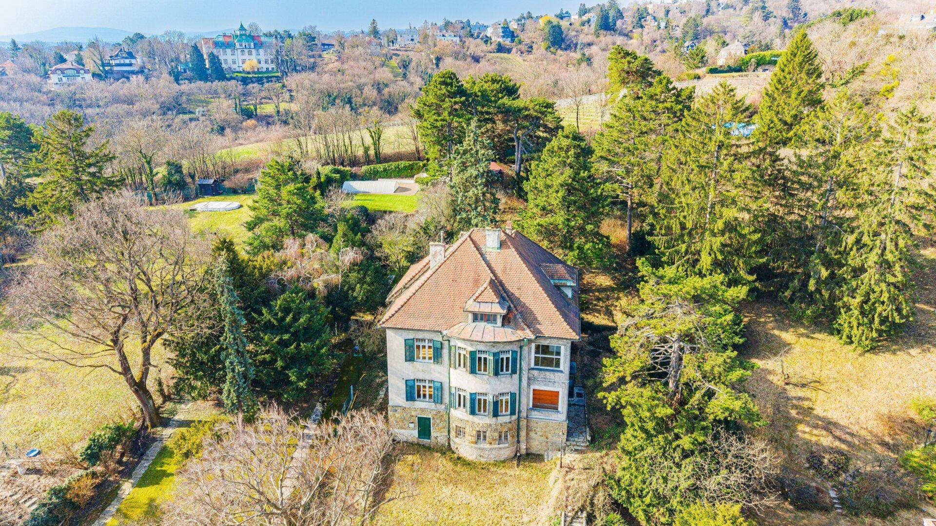 Eine historische Villa aus der Vogelperspektive, umgeben von Bäumen und einer hügeligen Landschaft.