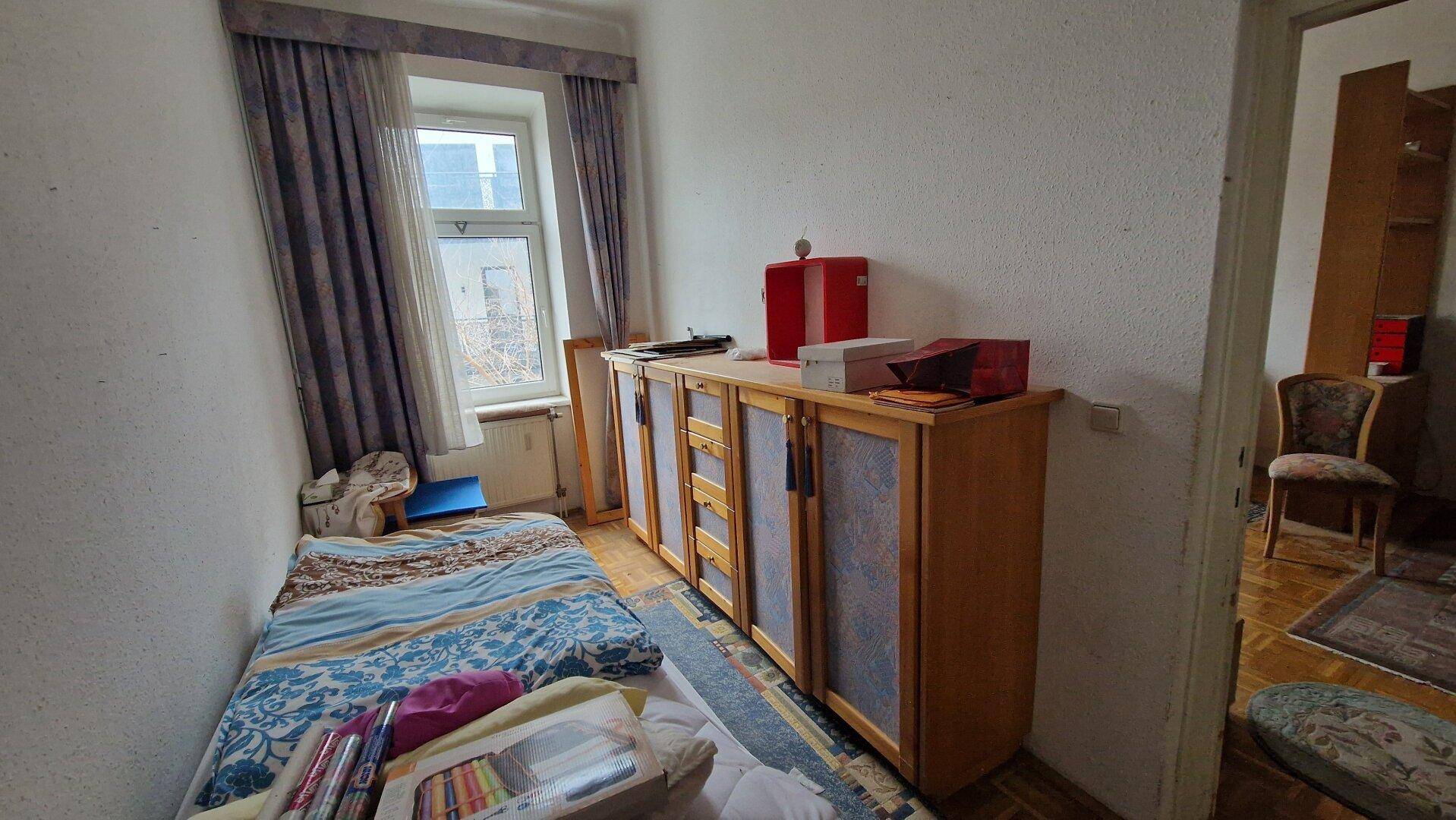 Kleines Schlafzimmer mit Einzelbett, Holzschrank und Fenster mit Vorhängen.