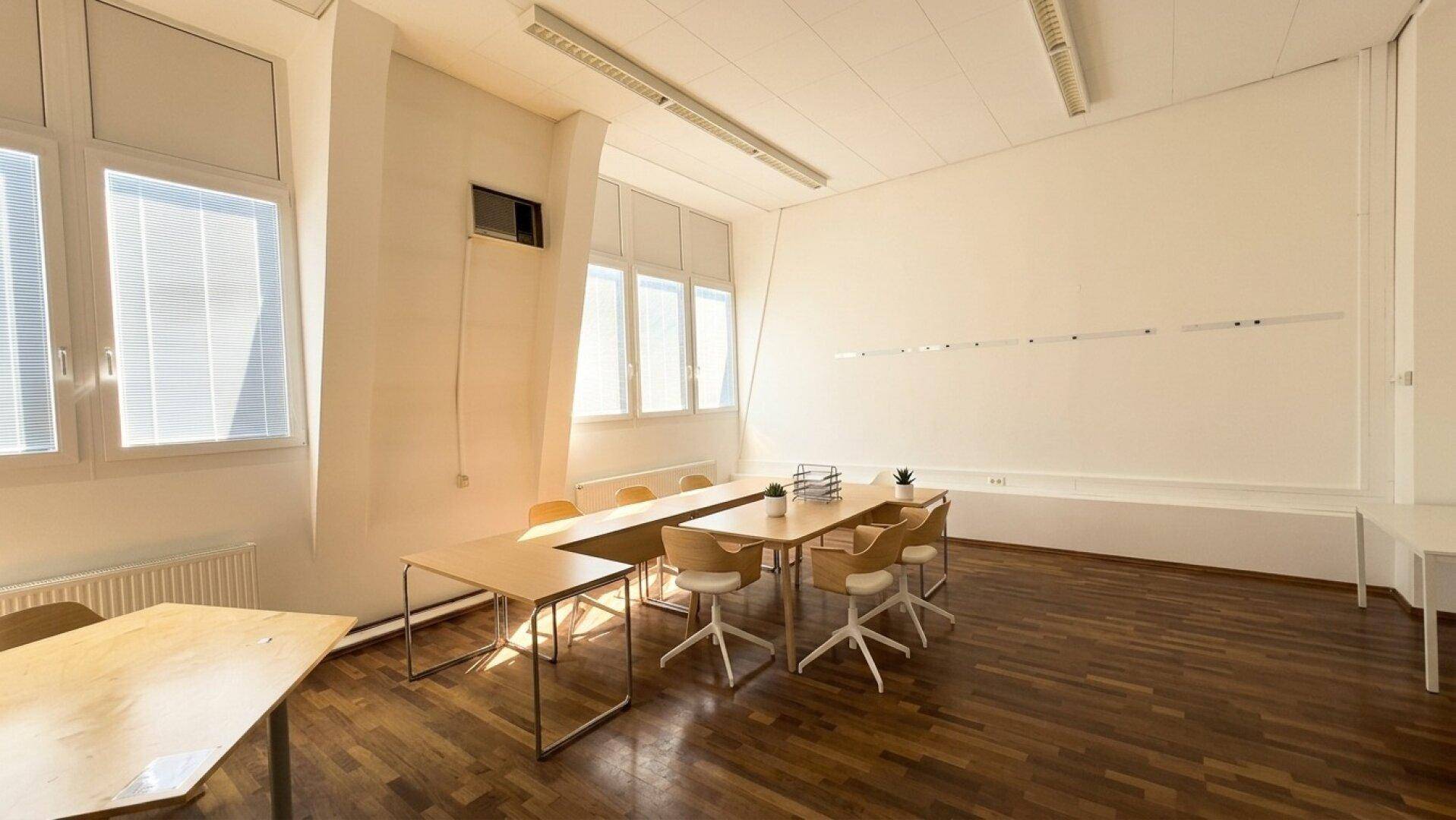 Geräumiger Schulungsraum mit Holzboden, Tischen und Stühlen, beleuchtet durch große Fenster.