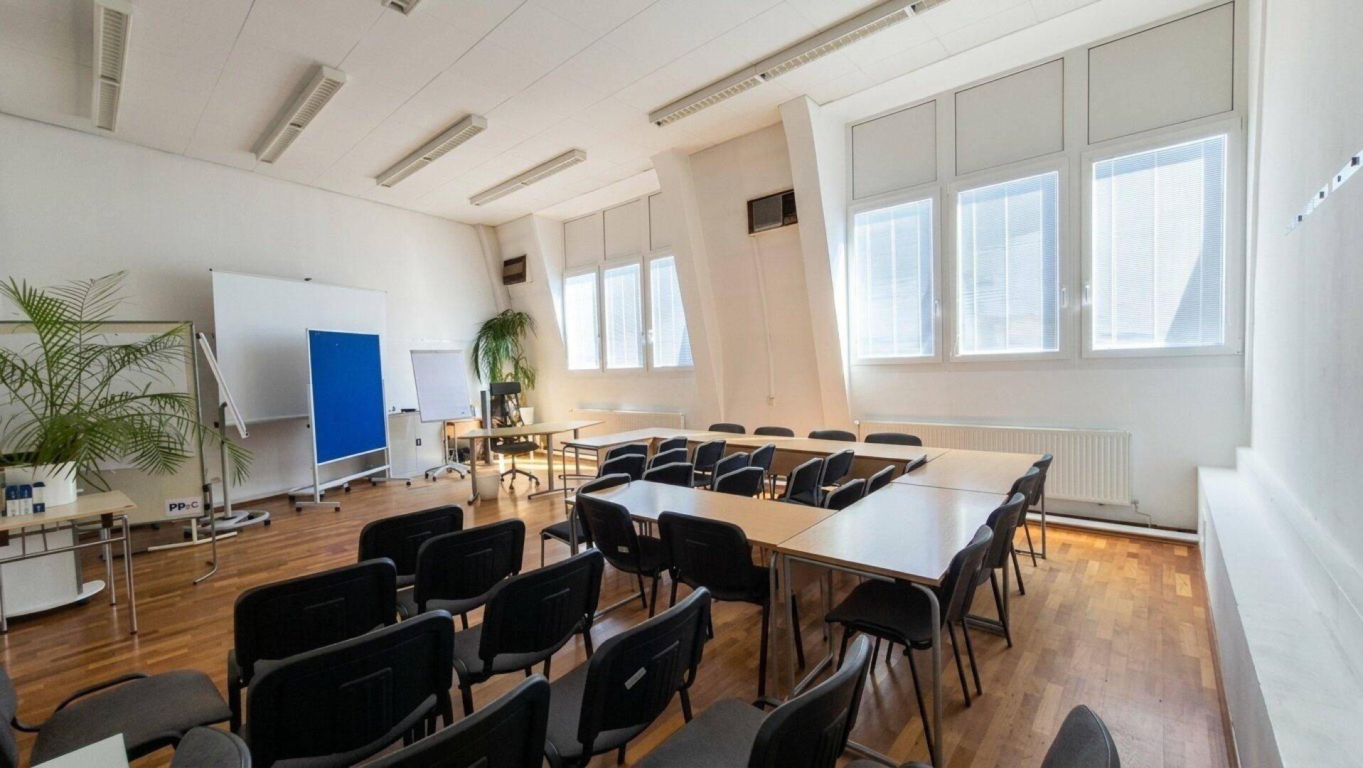 Heller Konferenzraum mit Holzboden, vielen Tischen und Stühlen sowie großen Fenstern.