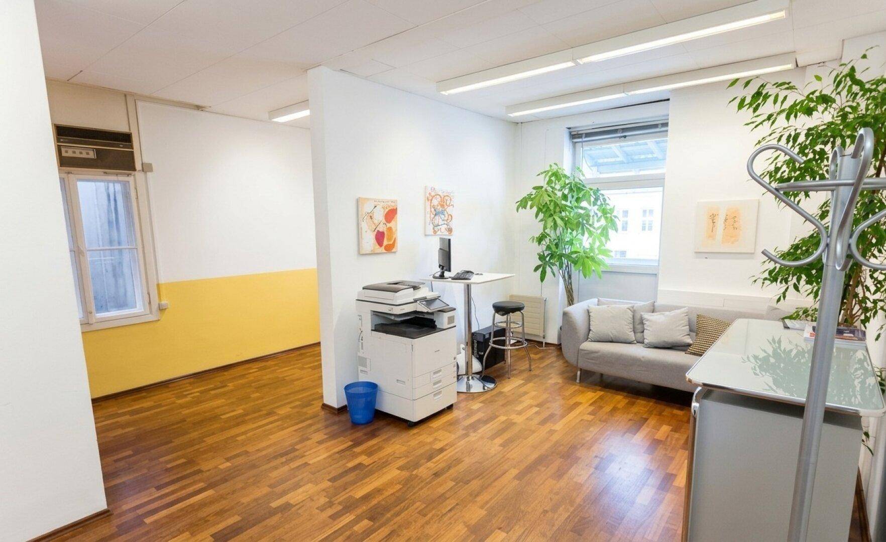 Empfangsbereich oder offenes Büro mit Holzboden, Sofa, Drucker und Pflanzen.