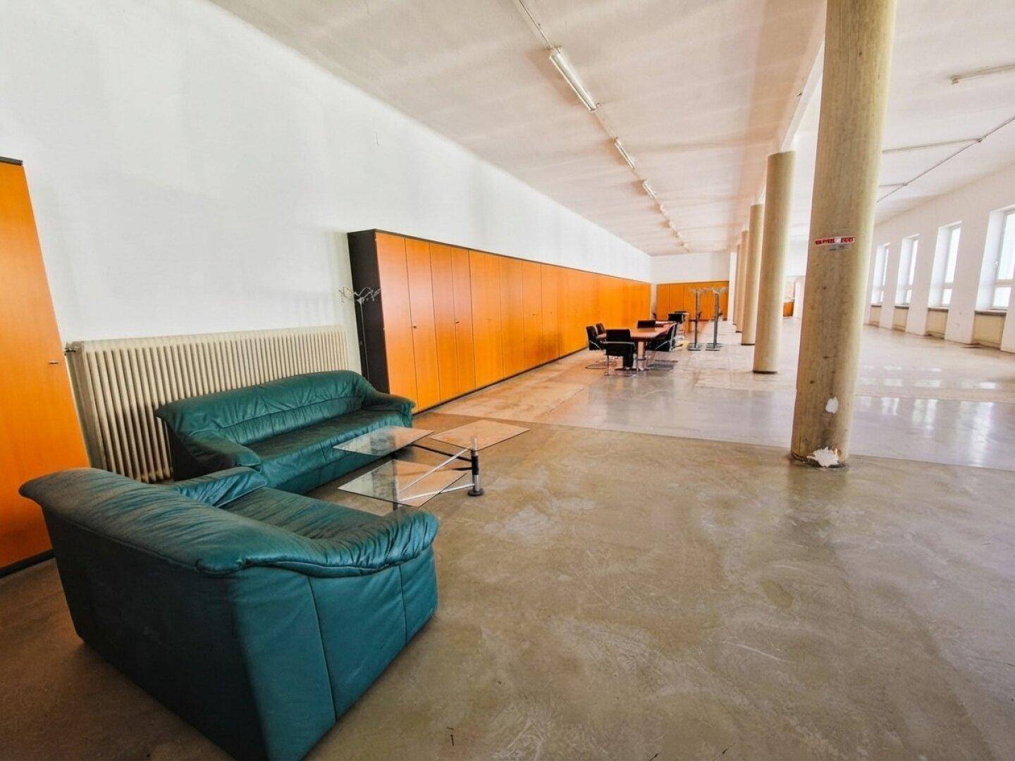 Weitläufiger Bürobereich mit grünen Sofas, Besprechungstisch und langen Holzschränken an der Wand, gut beleuchtet.