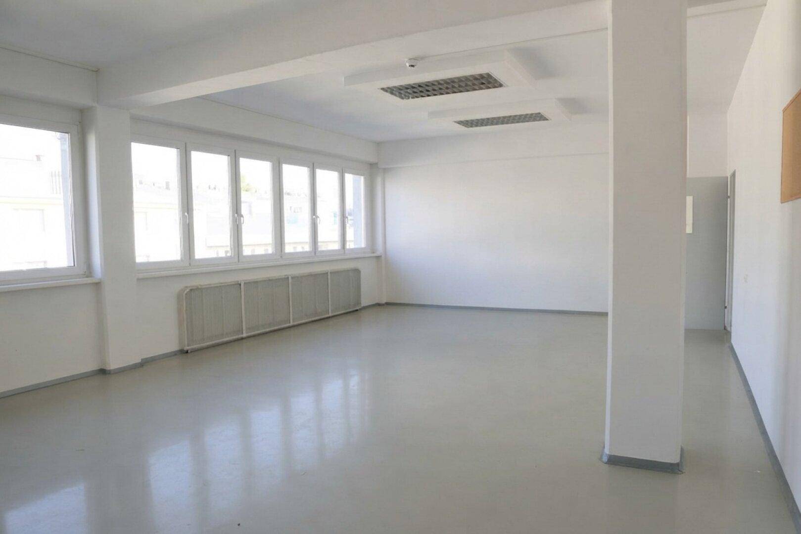Leerer Büroraum mit mehreren Fenstern, Heizkörpern unter den Fenstern und Deckenbeleuchtung.