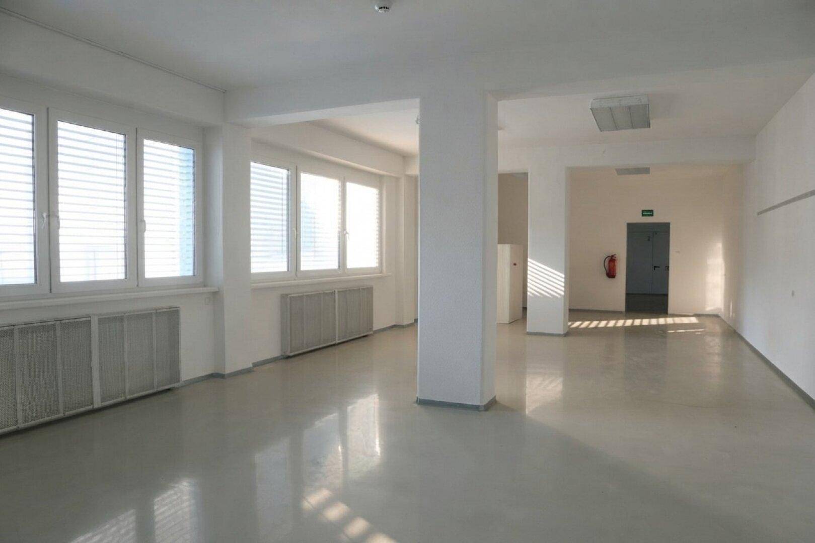 Heller, leerer Büroraum mit Fenstern, Jalousien und einem sichtbaren Notausgang.