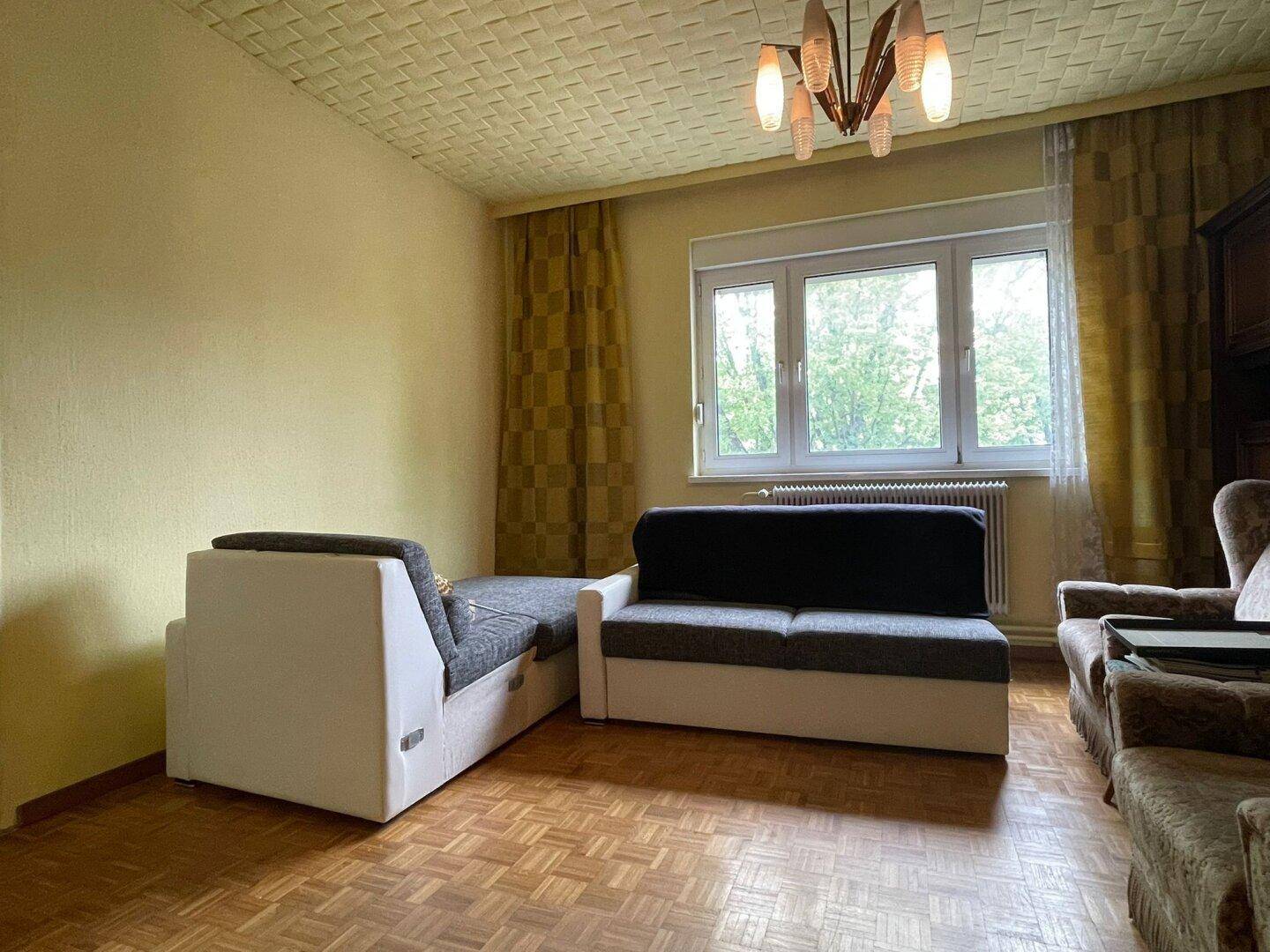 Wohnzimmer mit Parkettboden, gemusterter Decke und einem großen Fenster, das viel Tageslicht hereinlässt.
