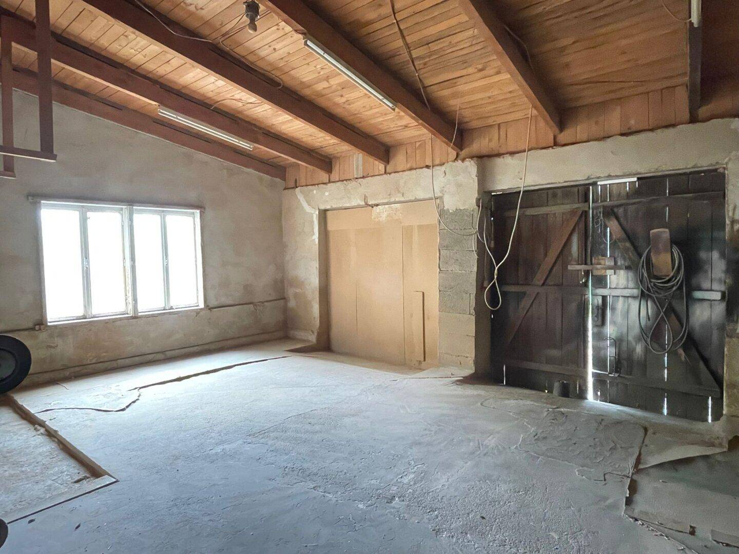 Großer, unfertiger Raum mit sichtbaren Holzbalken an der Decke und einem Betonboden, ideal für eine Werkstatt.