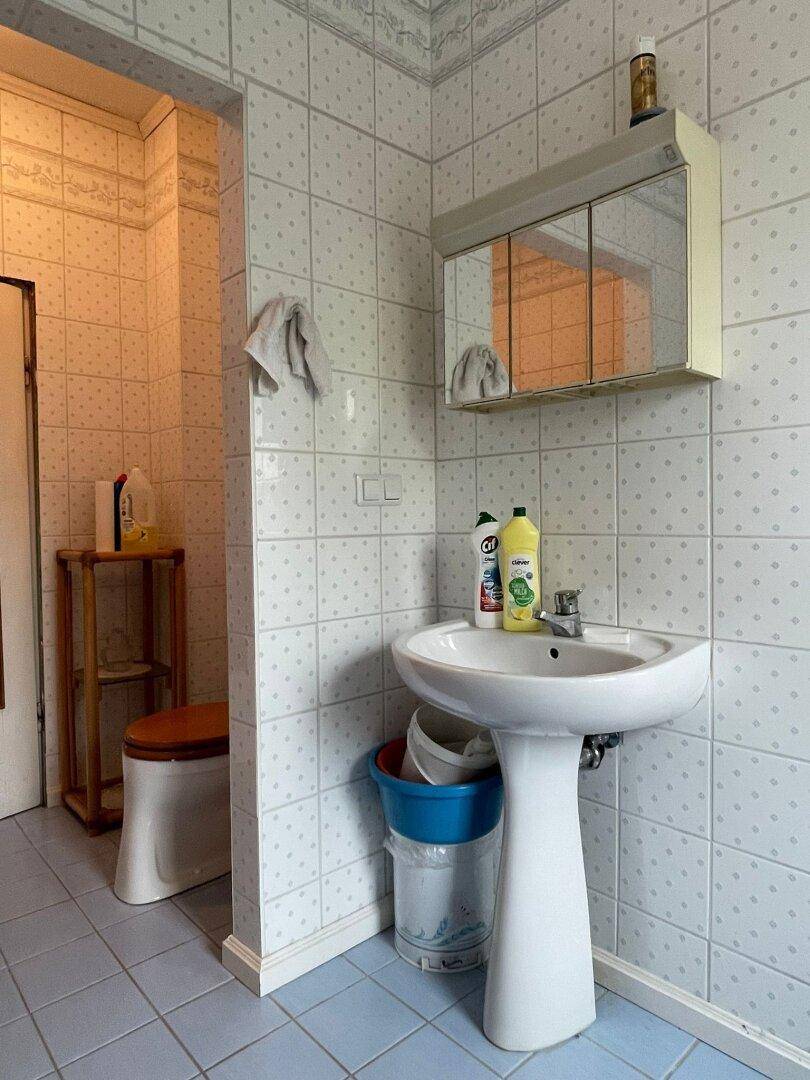 Badezimmerbereich mit Waschbecken, Spiegelschrank und hellblauen Fliesen, die einen älteren Stil aufweisen.