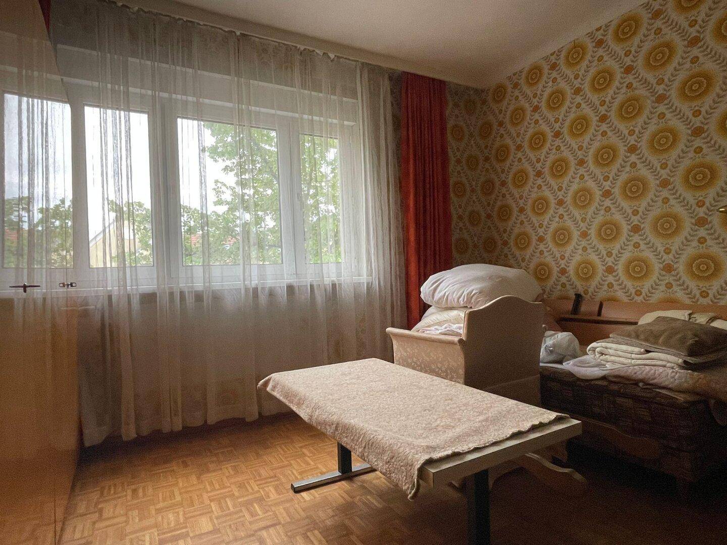 Schlafzimmer mit gemusterter Tapete, Parkettboden und einem großen Fenster, das den Raum erhellt.