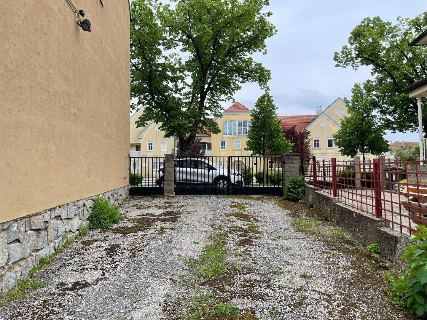 Unbefestigte Zufahrt oder Parkfläche neben dem Gebäude, umgeben von einer Mauer und Bäumen.