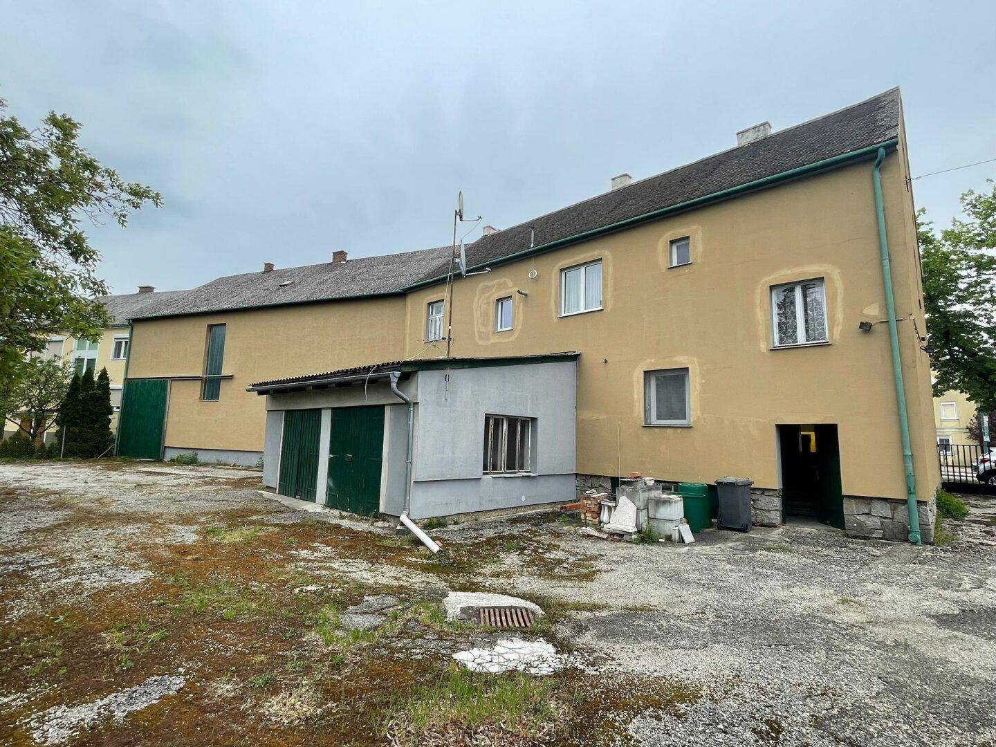 Rückseite eines älteren Gebäudes mit Anbau und Garagentoren, umgeben von einer unbefestigten Fläche.