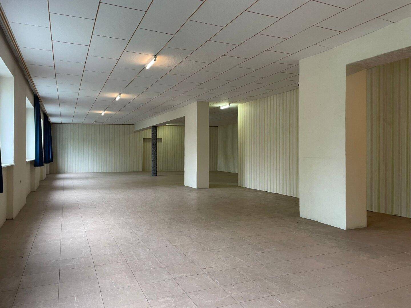 Weitläufiger, offener Raum mit Fliesenboden und mehreren Fenstern, der vielseitig nutzbar ist.