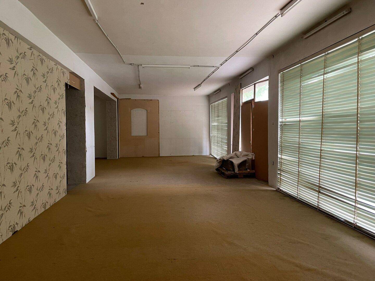 Großer, leerer Raum mit Teppichboden und Jalousien an den Fenstern, der Renovierung benötigt.