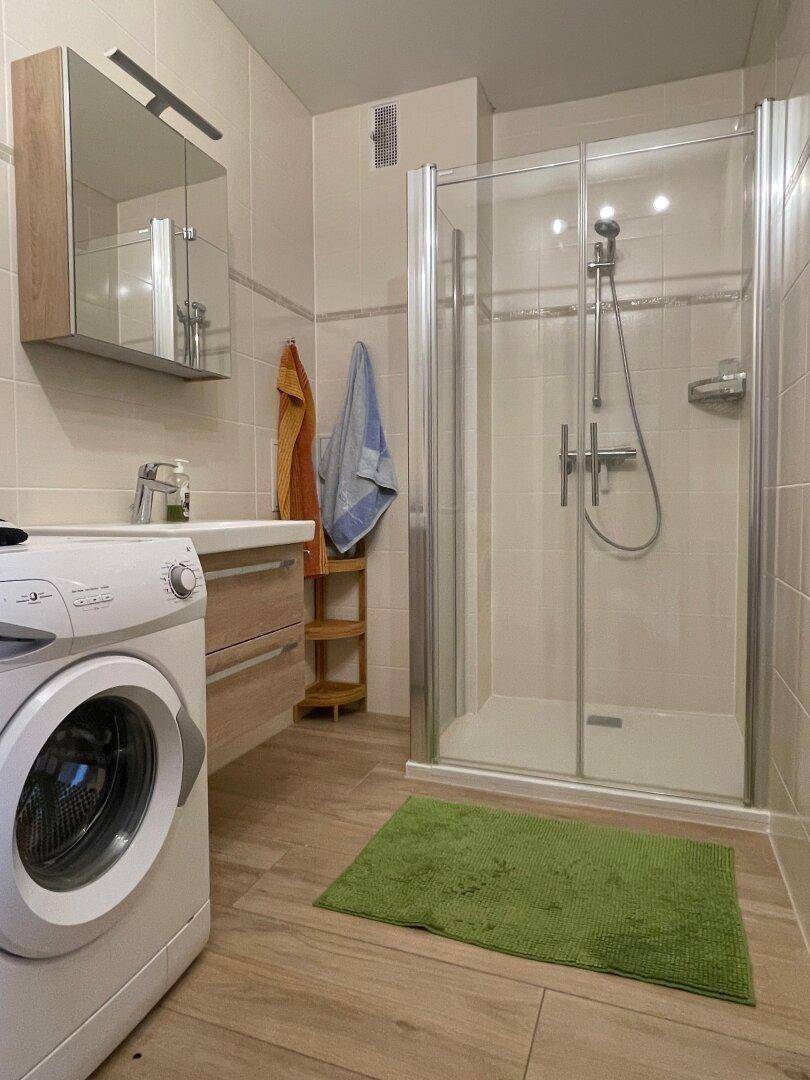 Modernes Badezimmer mit Duschkabine, Waschbecken und integrierter Waschmaschine, hell gefliest.