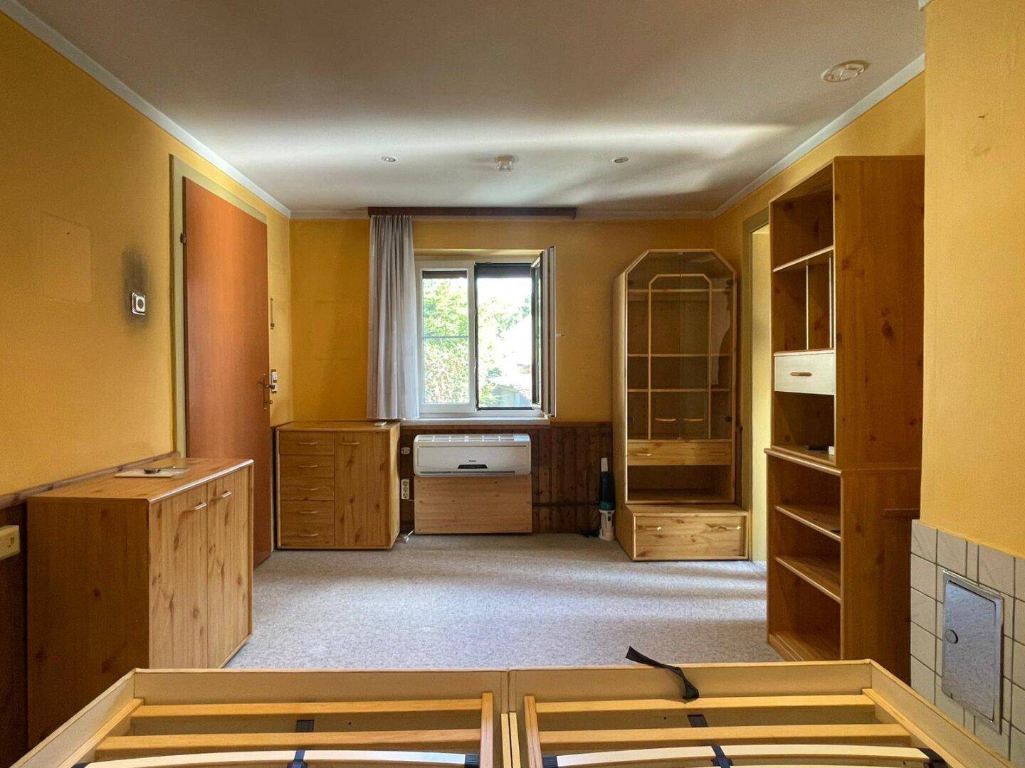 Schlafzimmer mit Teppichboden, Holzmöbeln und einem Fenster, das Tageslicht hereinlässt.