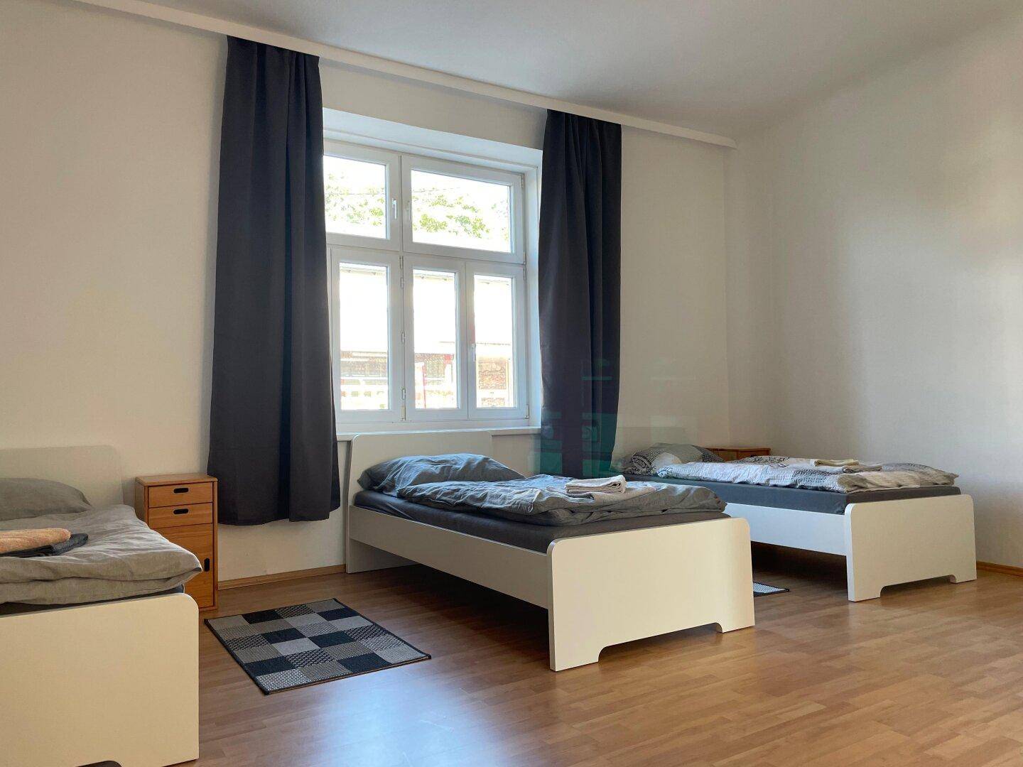 Geräumiges Schlafzimmer mit drei Betten, Holzboden und großen Fenstern.