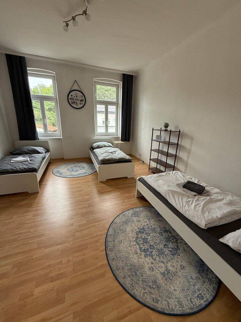 Helles Schlafzimmer mit drei Betten, Holzboden und zwei Fenstern.