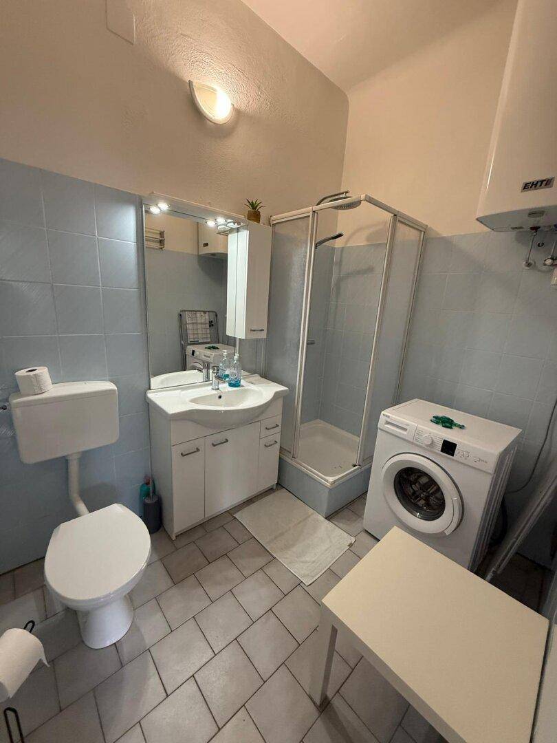 Badezimmer mit Dusche, WC, Waschbecken, Waschmaschine und hellblauen Fliesen.