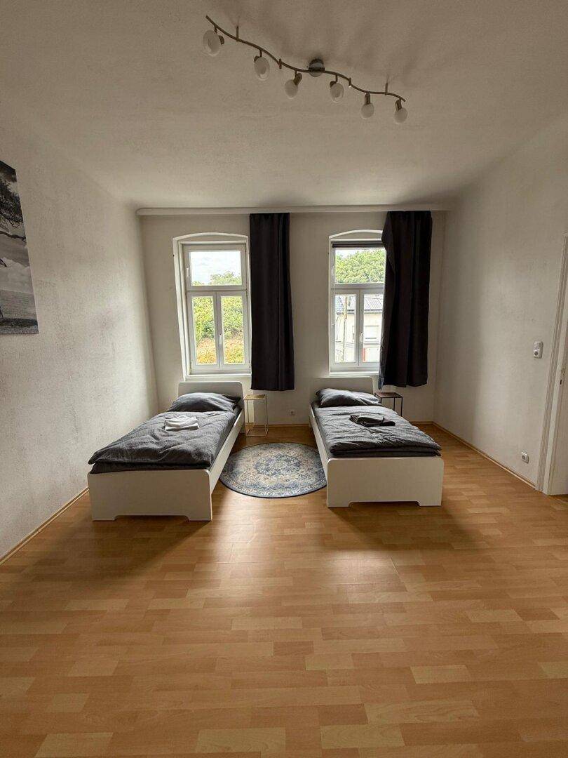 Helles Schlafzimmer mit zwei Betten, Holzboden und dunklen Vorhängen.