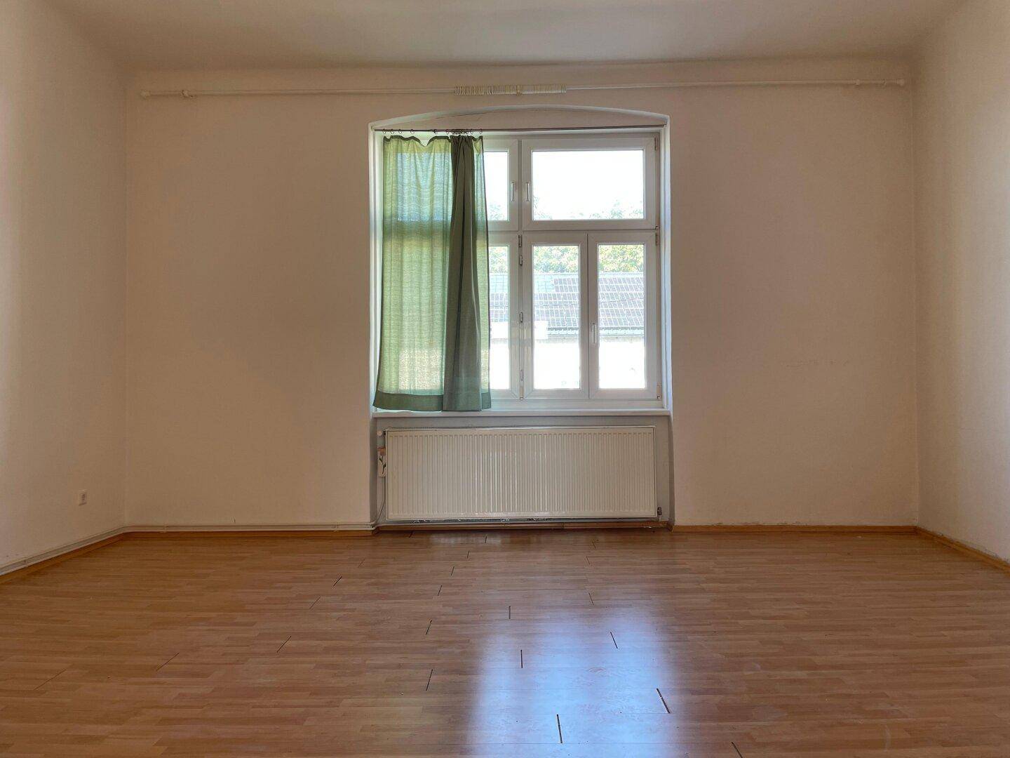 Leeres Wohnzimmer mit Holzboden, Fenster und grünen Vorhängen.