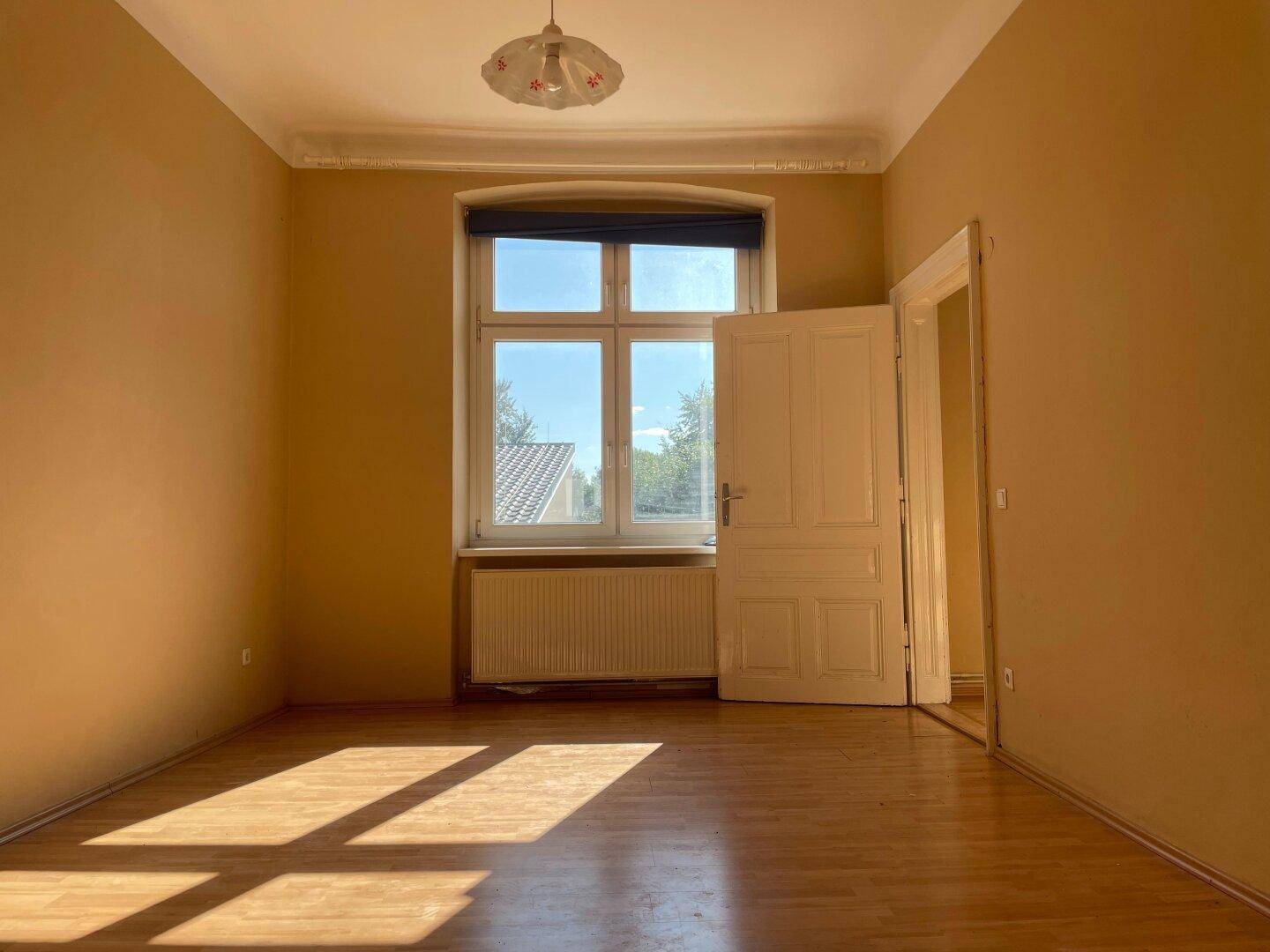 Leeres Wohnzimmer mit Holzboden, großem Fenster und viel Tageslicht.