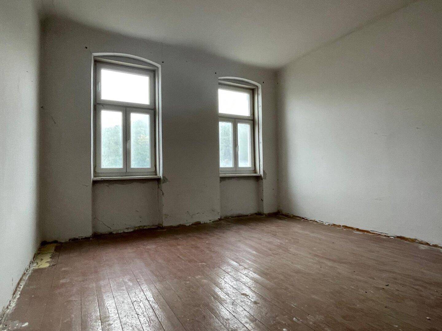 Leeres Schlafzimmer mit Holzboden und zwei Fenstern, renovierungsbedürftig.